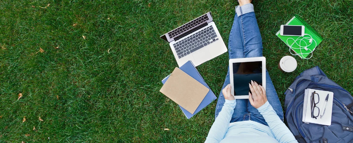 Sicht von oben: Eine Frau sitzt mit ausgestreckten Beinen auf einer Wiese. Sie arbeitet am Tablet, ein Note ist auf der Wiese, ebenso ein Rucksack, die Sonnebrille, ein Notizbuch und das Handy.