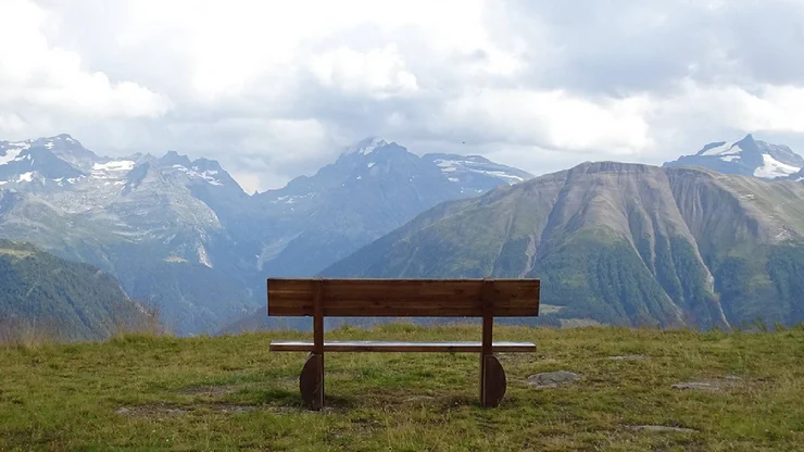 Eine Sitzbank auf einem Berg und dahinter das Bergpanorama und ein wolkiger Himmel.